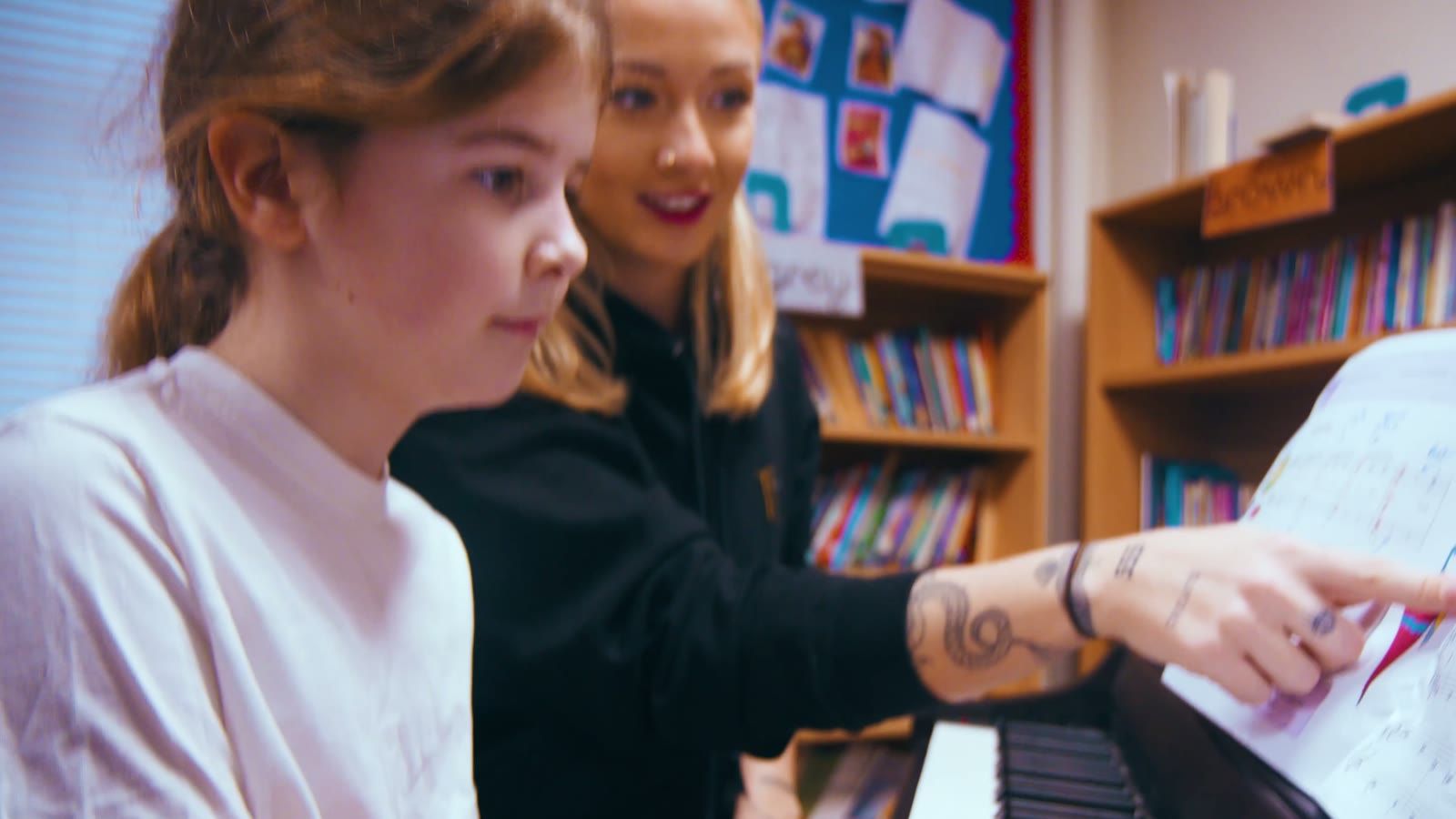 A music teacher smiles as a pupil concentrates at the piano during a lesson.