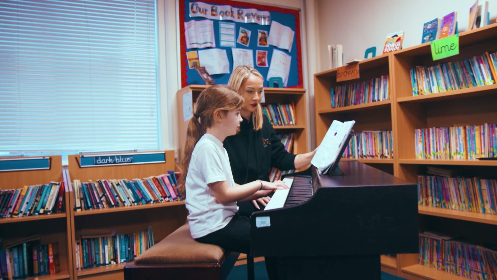 A music teacher sits beside a pupil at the piano in a primary school library, surrounded by bookshelves.